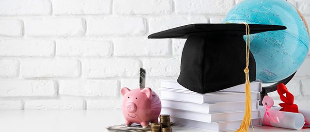 Graduation cap, piggy bank, and globe on a table, representing college and its costs.