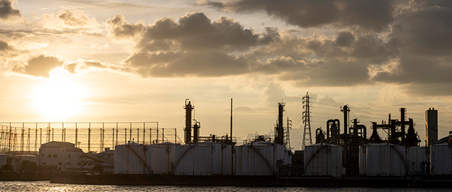 Oil storage tanks and refinery infrastructure along a waterfront at sunset, silhouetted against clouds and fading light. Licensed image by Freepick licensed to this website